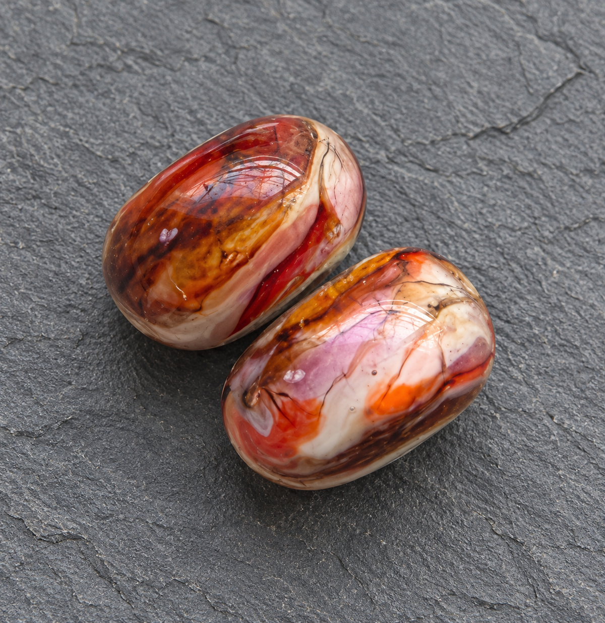 Two polished agate stone cabochons with swirling patterns of red, orange, brown, and cream, displayed on a dark gray slate surface.