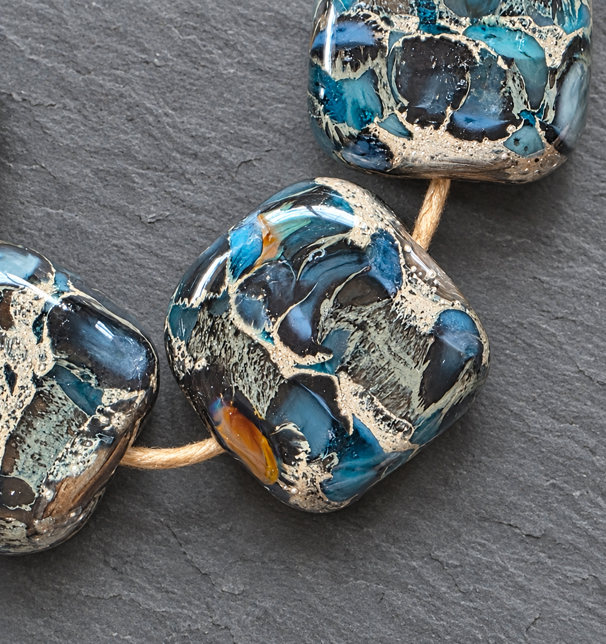 Close-up of handmade glass beads with a marbled pattern of blue, black, and metallic silver, threaded on a tan cord against a dark gray slate background.