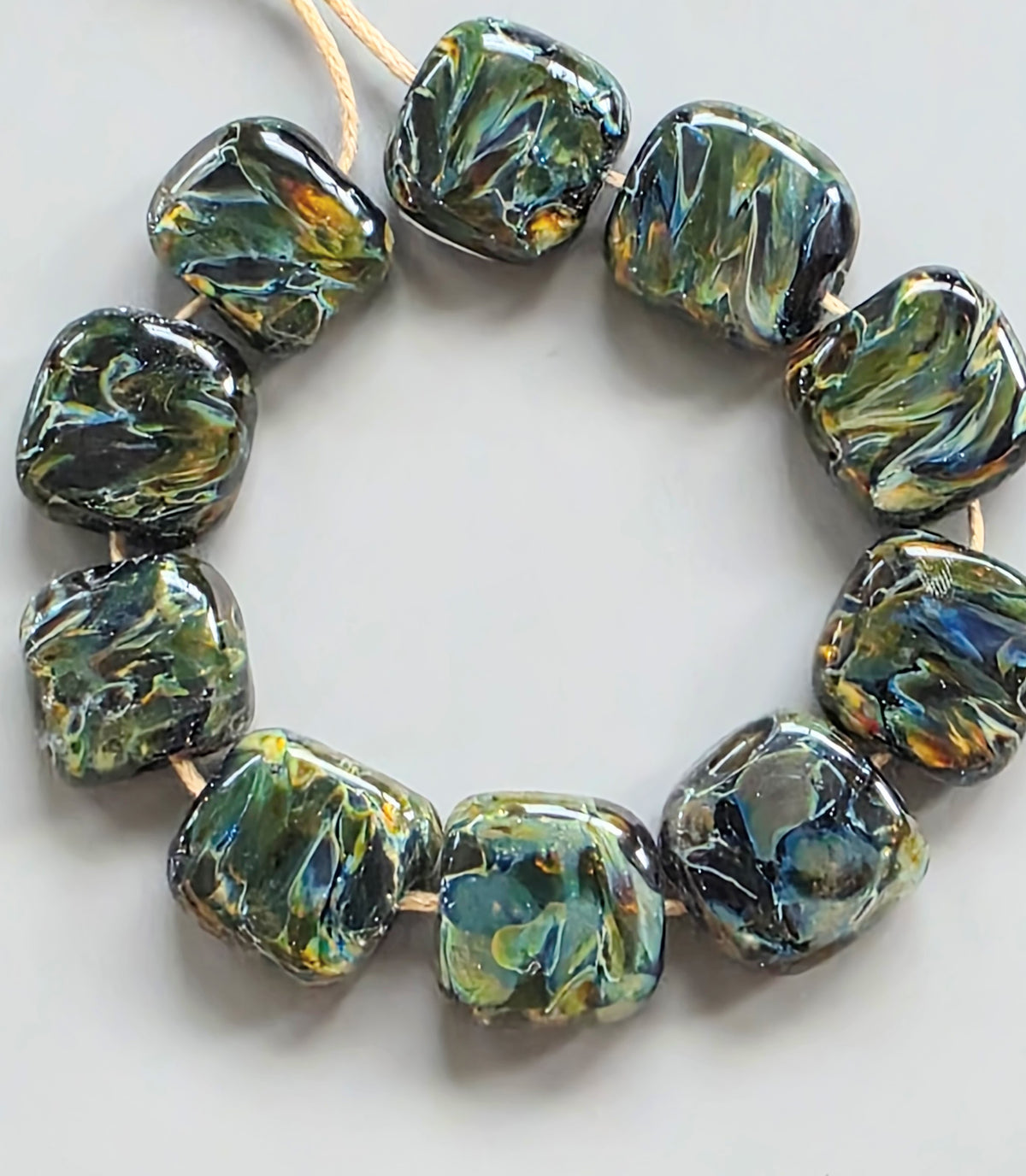 A strand of chunky, rounded square glass beads with a marbled swirl pattern in olive green, black, and iridescent blue, strung on a light tan cord against a neutral light gray background.