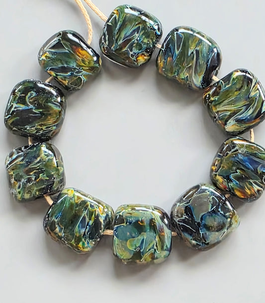 A strand of chunky, rounded square glass beads with a marbled swirl pattern in olive green, black, and iridescent blue, strung on a light tan cord against a neutral light gray background.