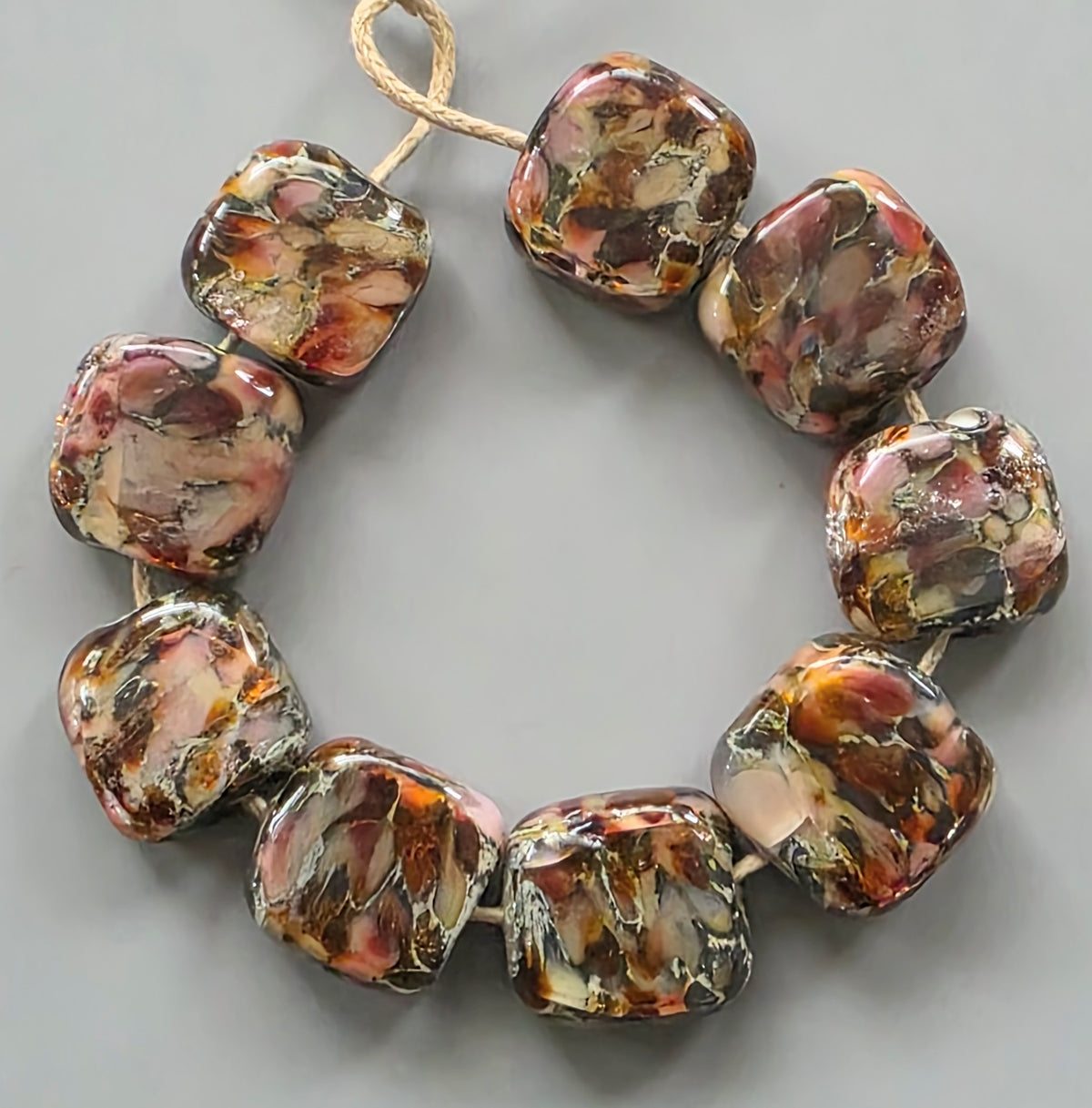 A strand of chunky, rounded square glass beads with a marbled pattern of brown, orange, pink, and cream, strung on a natural jute cord against a neutral gray background.