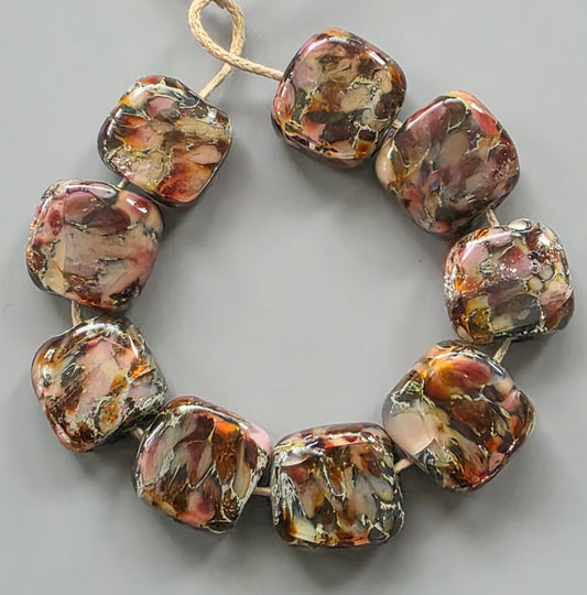A strand of chunky, rounded square glass beads with a marbled pattern of brown, orange, pink, and cream, strung on a natural jute cord against a neutral gray background.