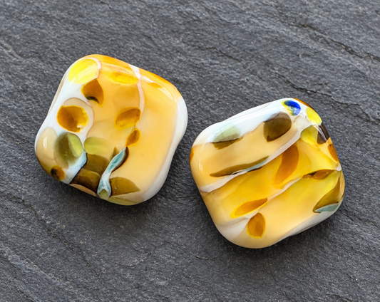 Two handmade glass lampwork beads with a marbled pattern of yellow, white, olive green, and light blue, displayed on a dark gray slate background.