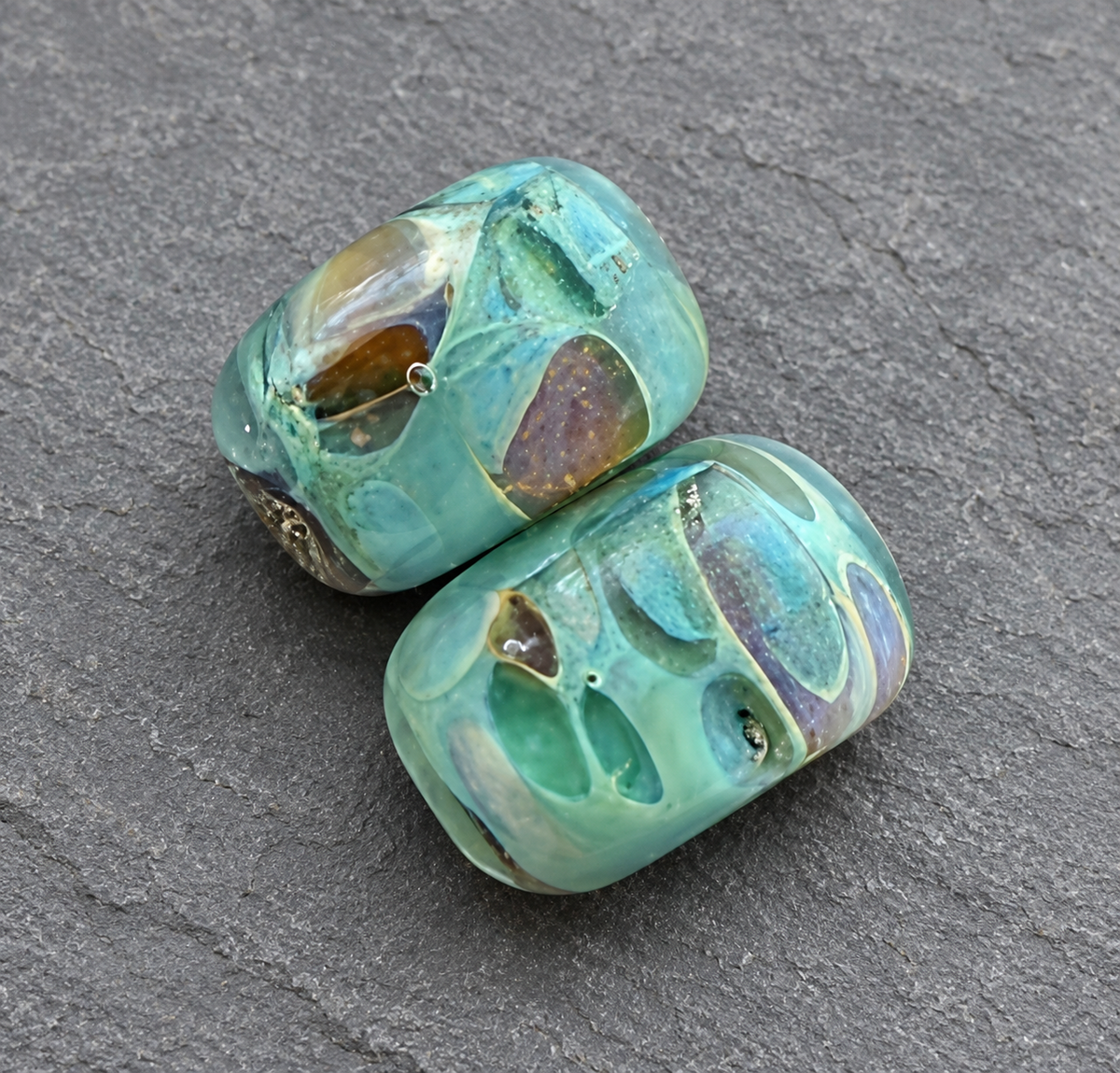 Two handmade glass lampwork beads with a marbled pattern of turquoise, green, and iridescent blue, resting on a dark gray slate surface.