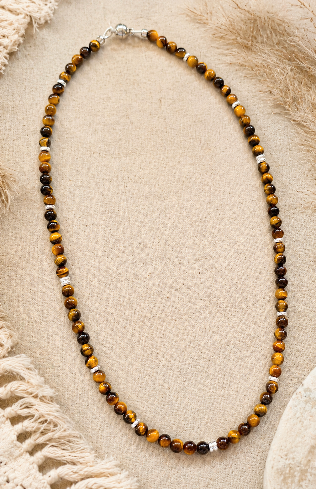 A beaded necklace made of alternating brown and black tiger's eye beads with small silver spacer beads, displayed on a textured beige fabric background.