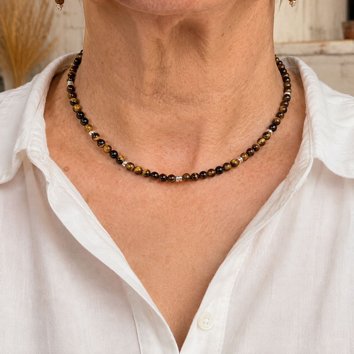 A close-up of a woman's neck and upper chest wearing a beaded necklace made of dark brown, black, and metallic gold beads, paired with a white collared shirt.