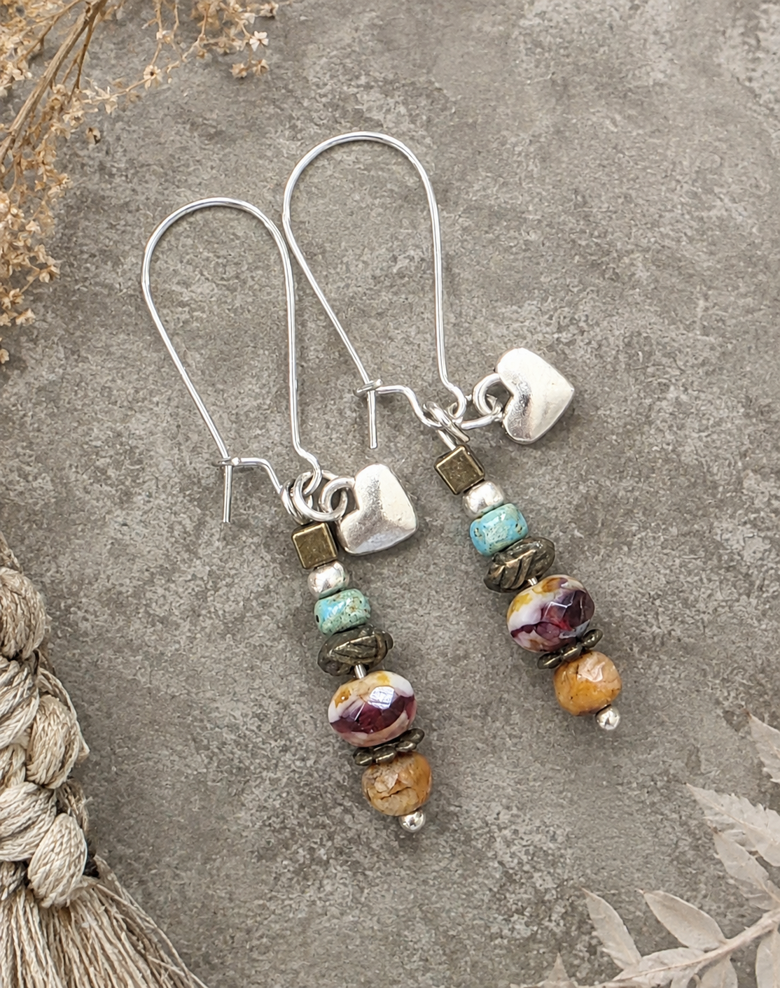 A pair of handmade dangle earrings with silver fishhook wires, featuring a small white heart charm, turquoise and bronze spacer beads, and marbled glass beads in purple, orange, and brown, displayed on a textured gray stone surface.