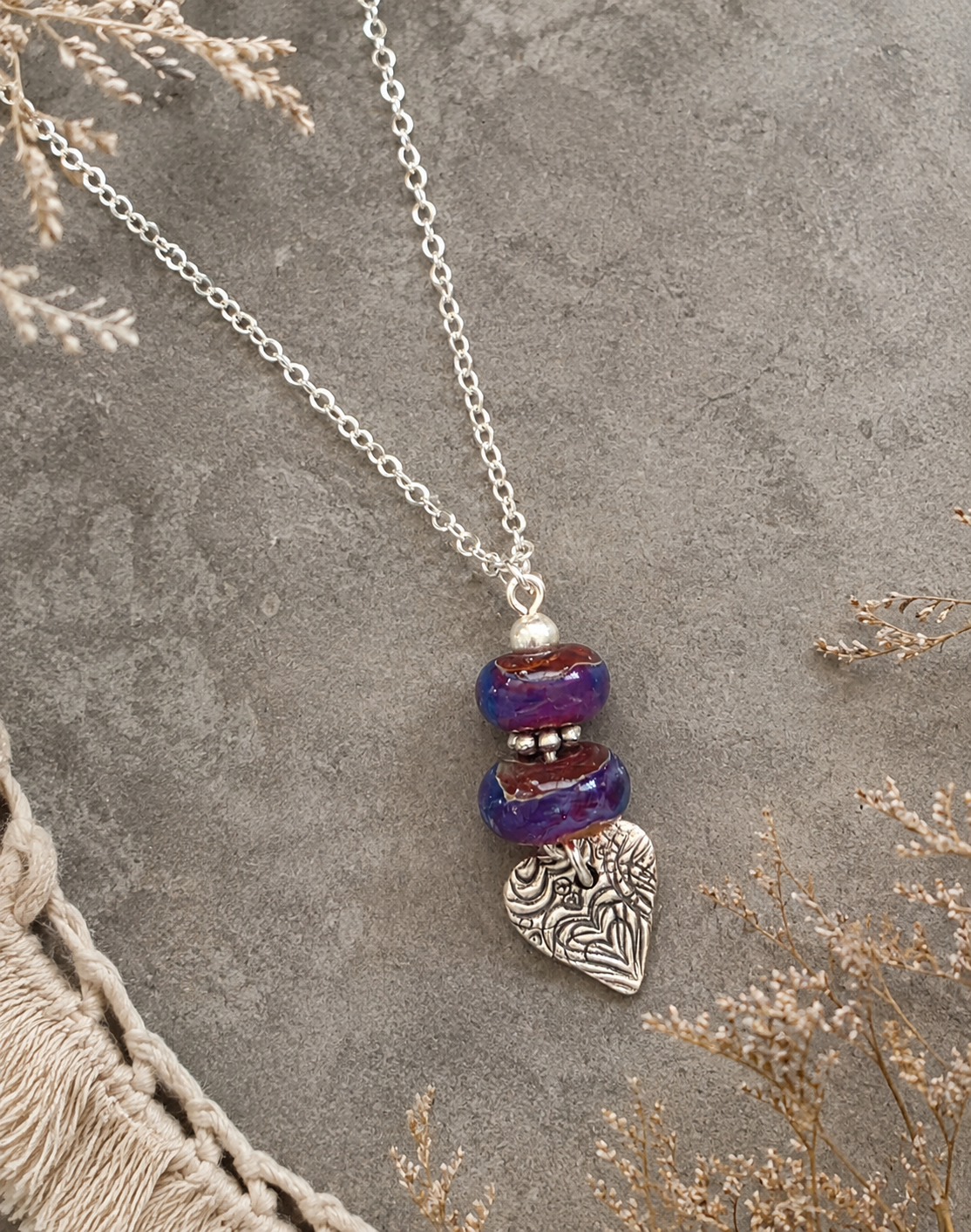 A silver chain necklace with a bohemian-style pendant featuring three stacked purple and red marbled beads and a silver heart-shaped charm with an engraved tree of life pattern, displayed on a textured gray surface with dried floral accents.