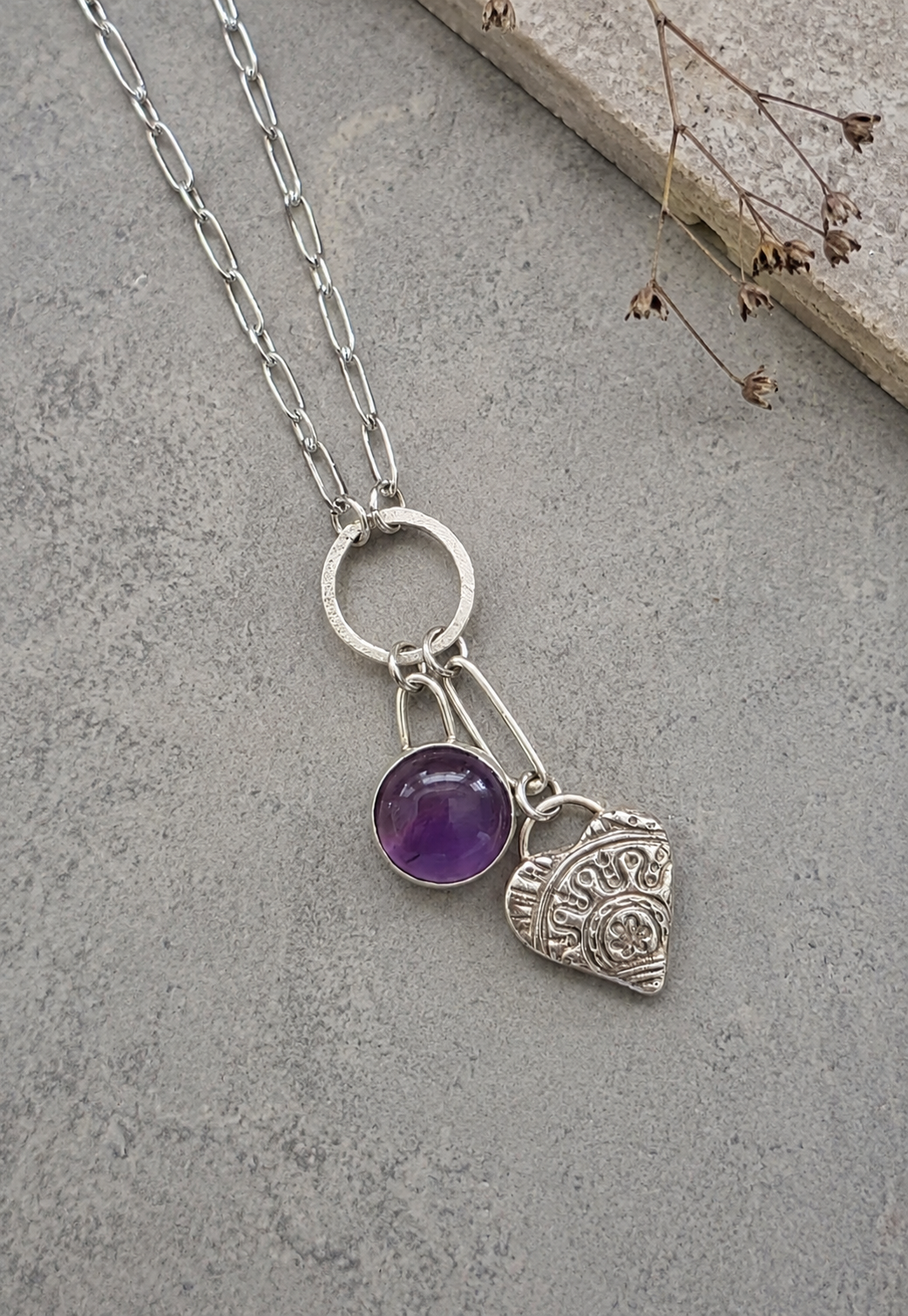 A silver-tone chain necklace with a circular ring connector holding a round purple amethyst gemstone and an engraved silver heart charm, displayed on a textured gray surface.