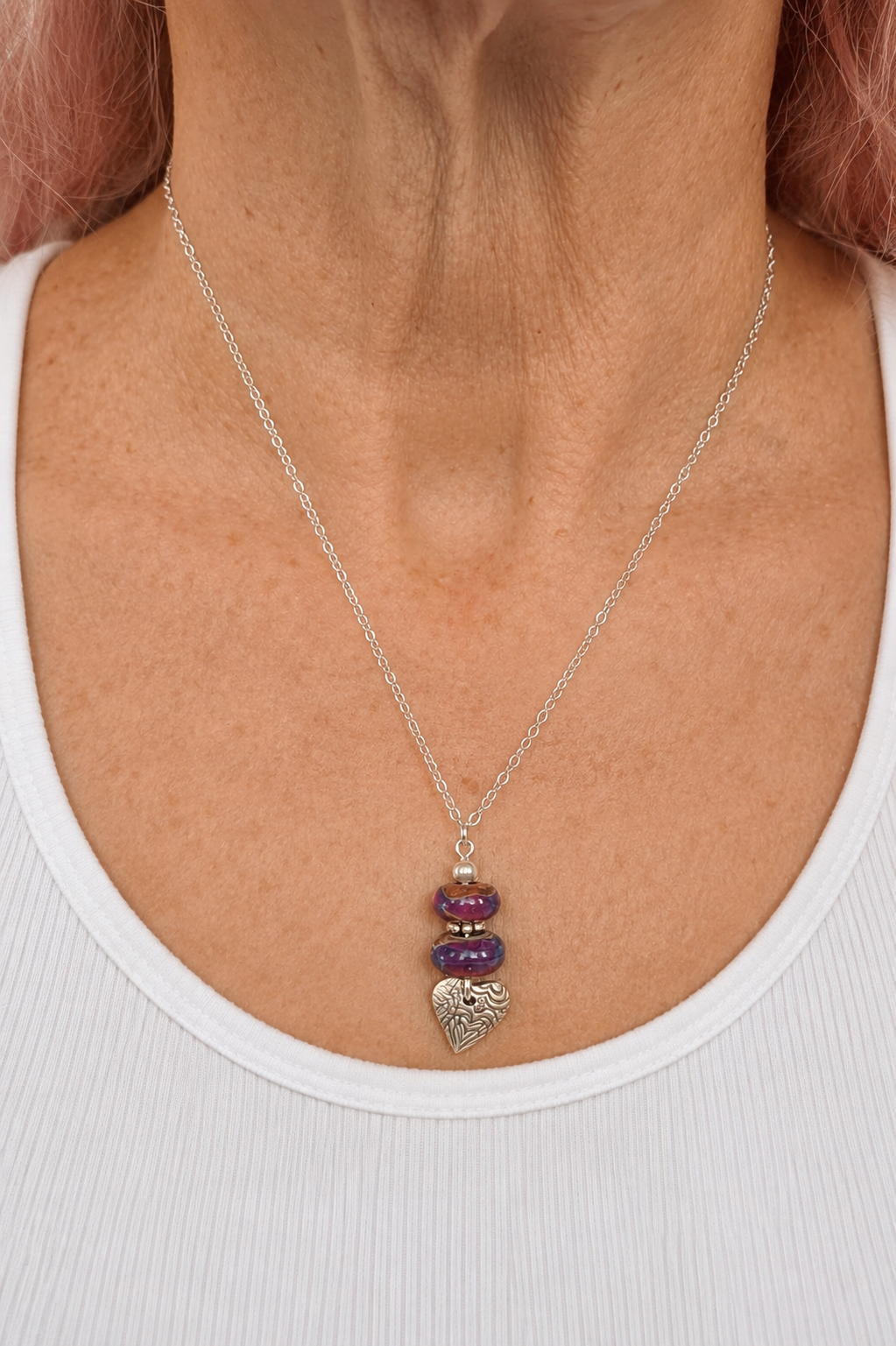 A close-up of a model wearing a delicate silver chain necklace with a triple-stone pendant featuring purple amethyst beads and a textured silver heart.