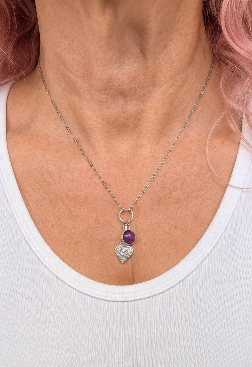 A close-up of a person wearing a delicate silver chain necklace with a small purple bead and a silver heart-shaped charm, styled with a white ribbed tank top.