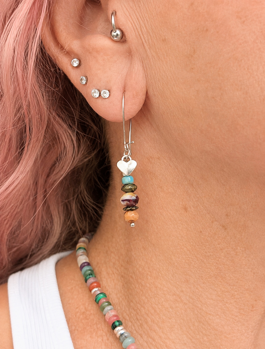A close-up of a person's ear wearing a silver-toned dangle earring with a white heart-shaped charm and stacked multicolored beads, alongside a matching beaded necklace and pink hair.