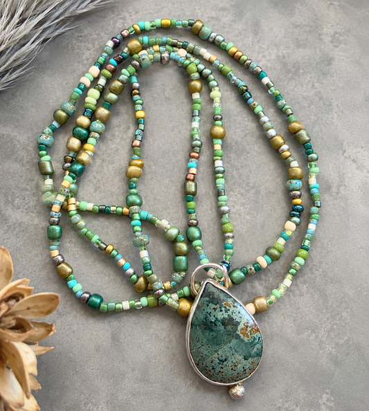 A beaded necklace with multiple strands of green, turquoise, and gold-toned beads, featuring a large teardrop-shaped green stone pendant with a silver-toned bezel, displayed on a textured gray surface with dried floral accents.