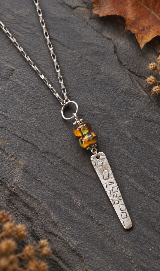A silver pendant necklace featuring a textured rectangular bar and a small amber glass bead cluster, displayed on a dark slate surface with dried leaves.