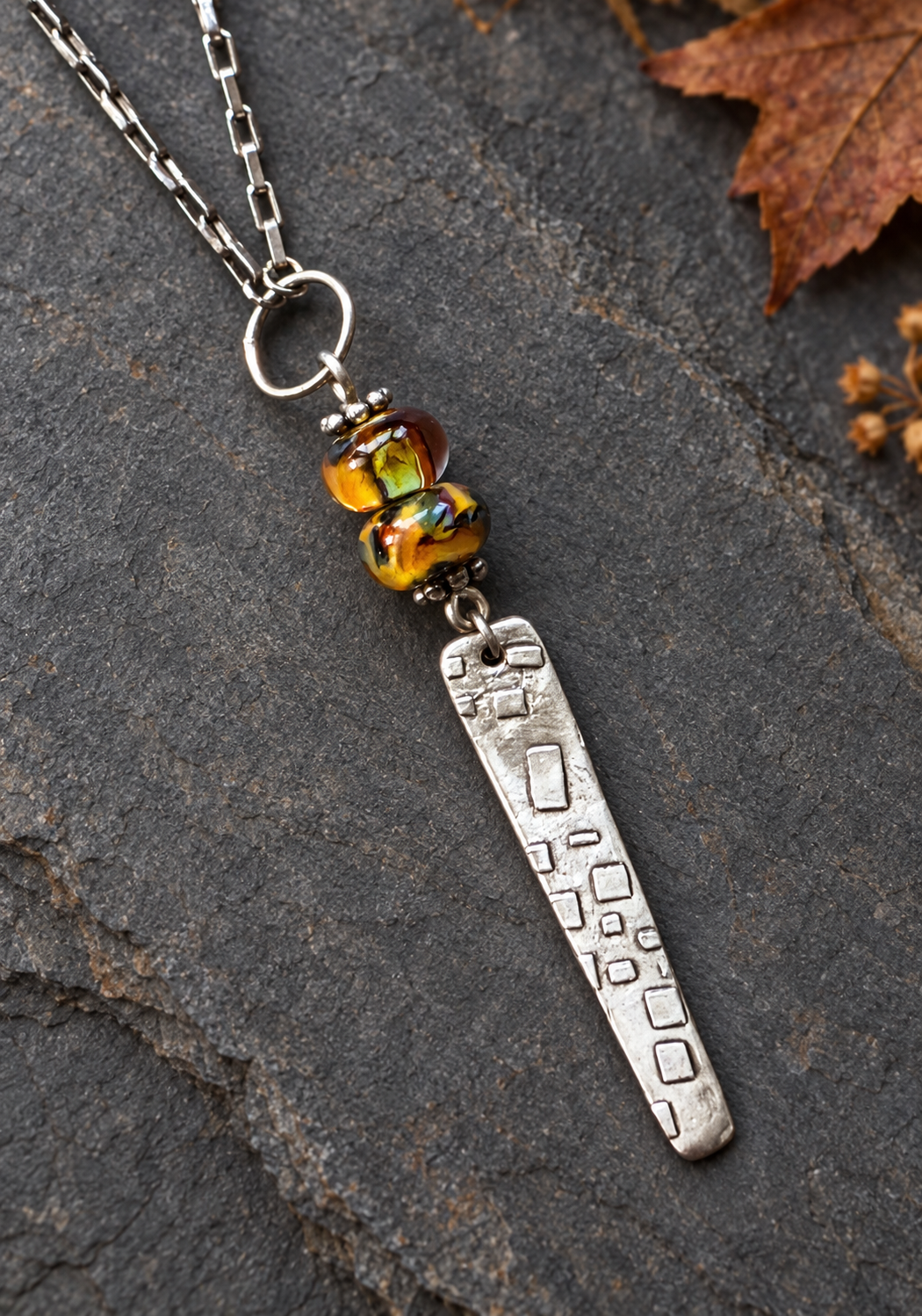 A silver-toned pendant necklace with a textured rectangular bar charm, a small round bead, and a colorful amber glass bead, displayed on a dark stone surface with a dry leaf.