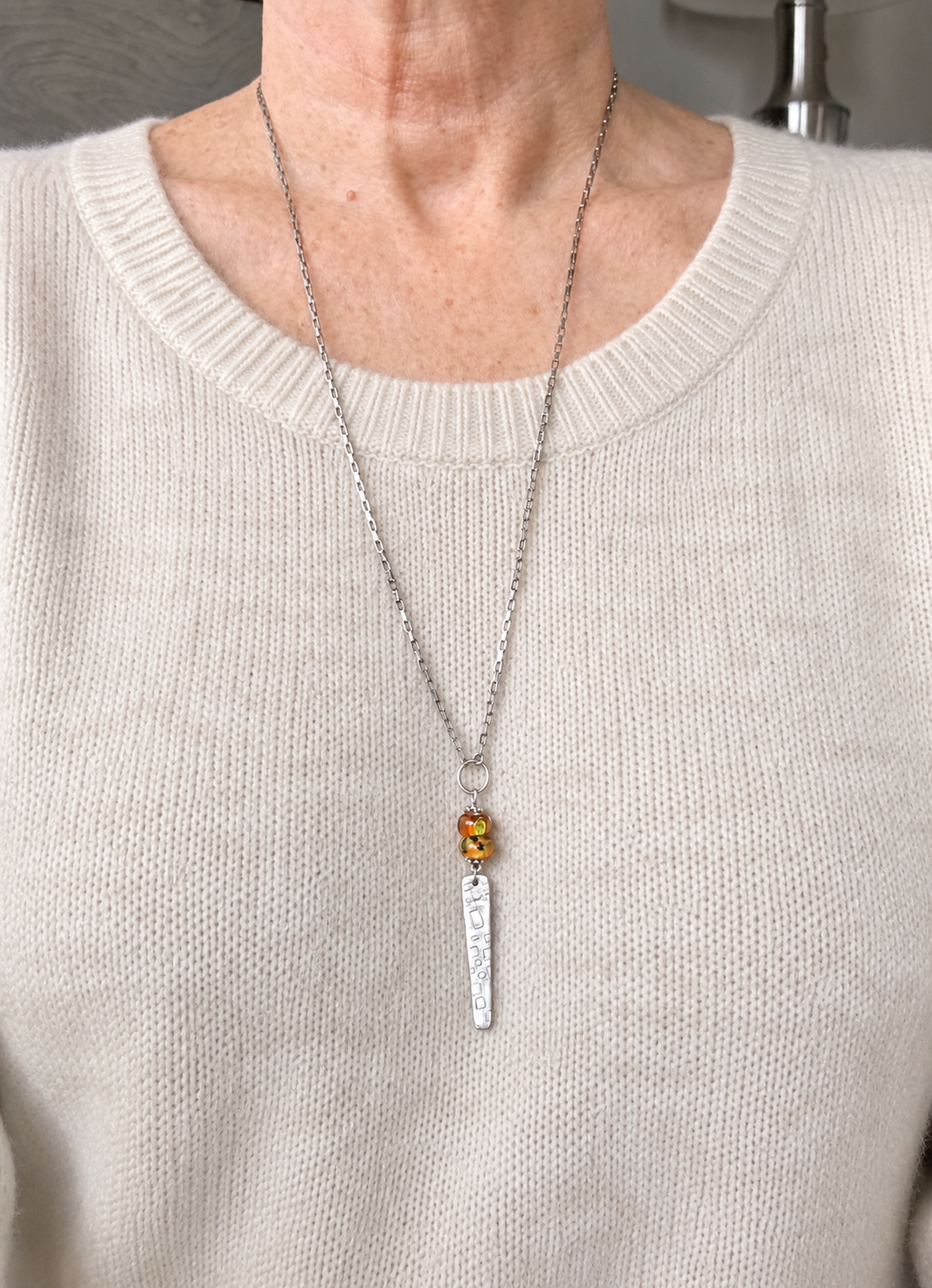 A close-up of a person wearing a silver necklace with a small amber-colored bead and a vertical engraved silver pendant, paired with a cream-colored knit sweater.