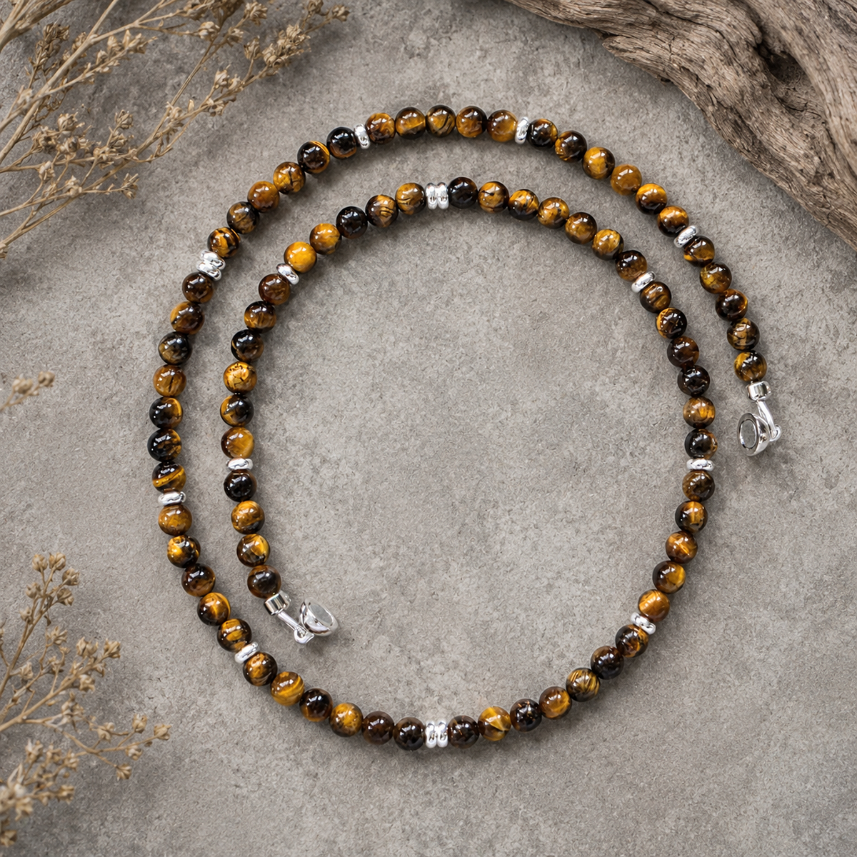 A beaded necklace made of round brown and black tiger's eye beads with small silver spacer beads and a silver clasp, arranged on a textured gray surface with dried floral props.