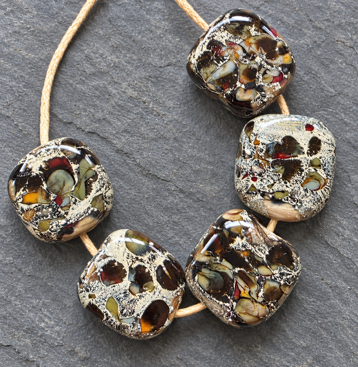 Five handmade square glass lampwork beads with a marbled pattern of black, brown, gold, and red, threaded onto a natural tan cord against a dark gray slate background.
