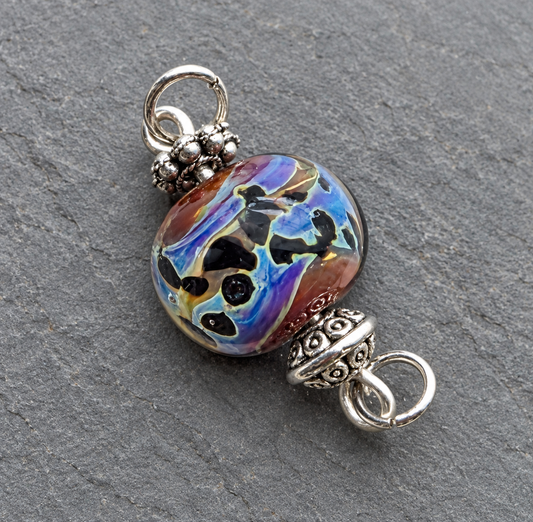 A round glass lampwork bead with a multicolor swirl pattern of purple, blue, black, and brown, set in silver-toned metal with decorative filigree spacers, shown on a textured gray slate background.