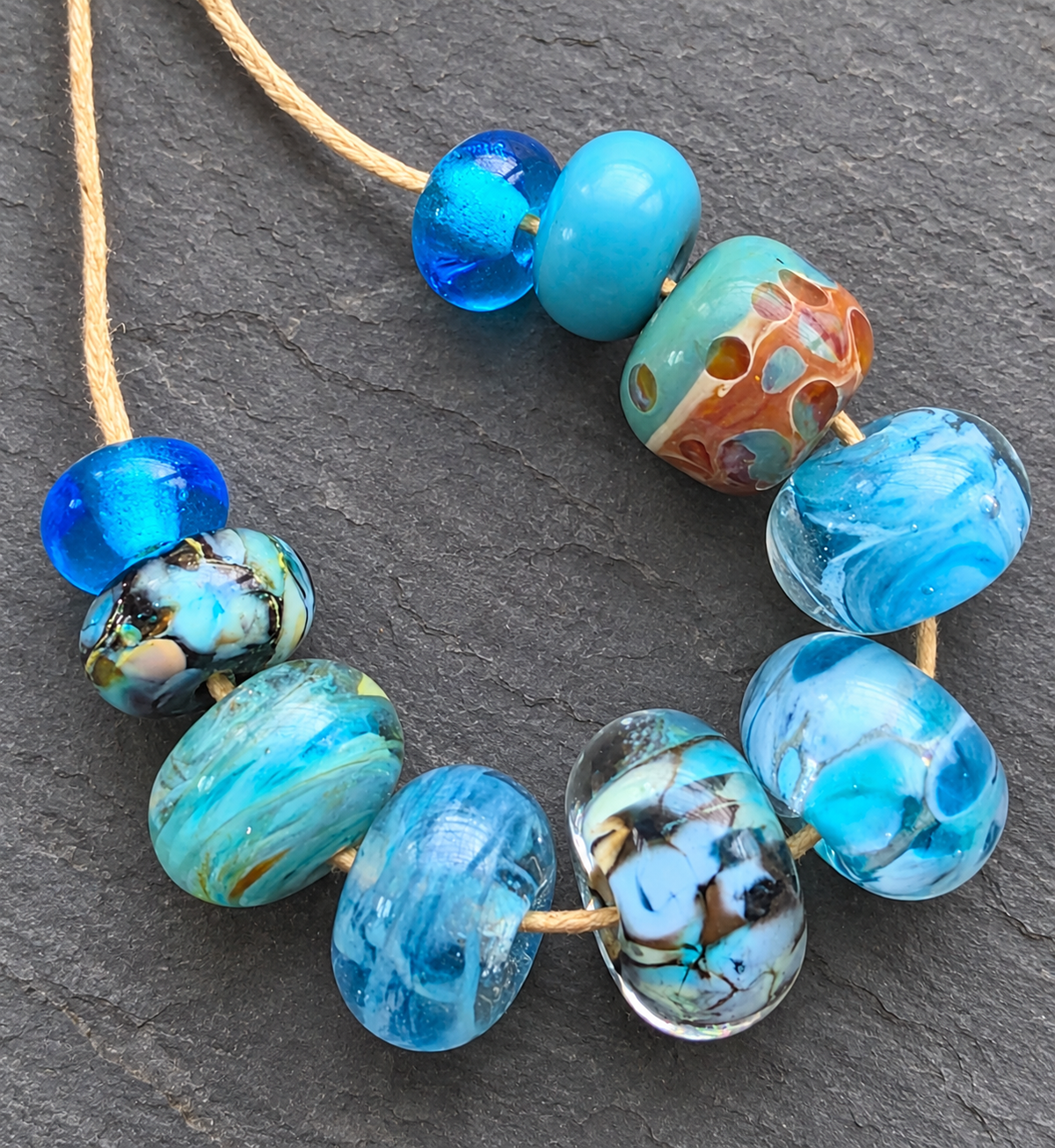 A strand of handmade glass lampwork beads in shades of blue, turquoise, and marbled patterns, threaded on a natural tan cord against a dark gray slate background.