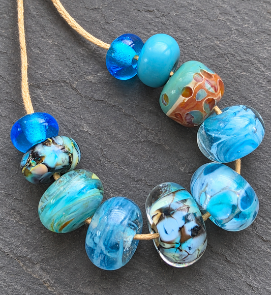 A strand of handmade glass lampwork beads in shades of blue, turquoise, and marbled patterns, threaded on a natural tan cord against a dark gray slate background.