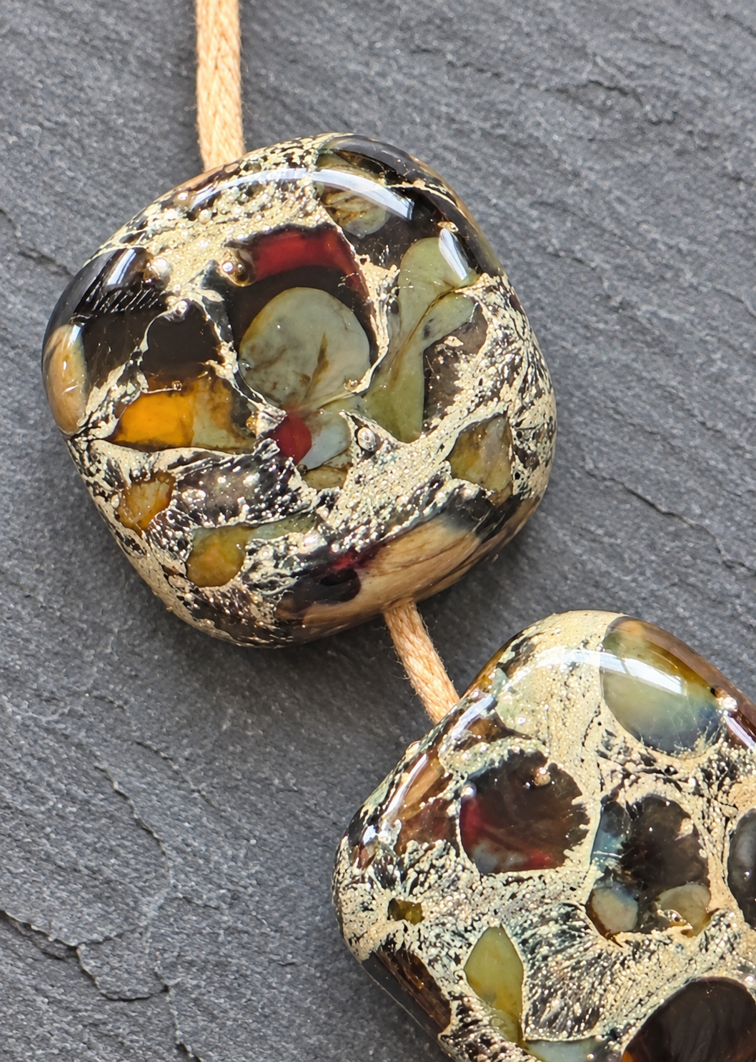Two handmade glass lampwork beads with a textured gold foil pattern and embedded multicolored glass pieces, threaded on a tan cord against a dark slate background.