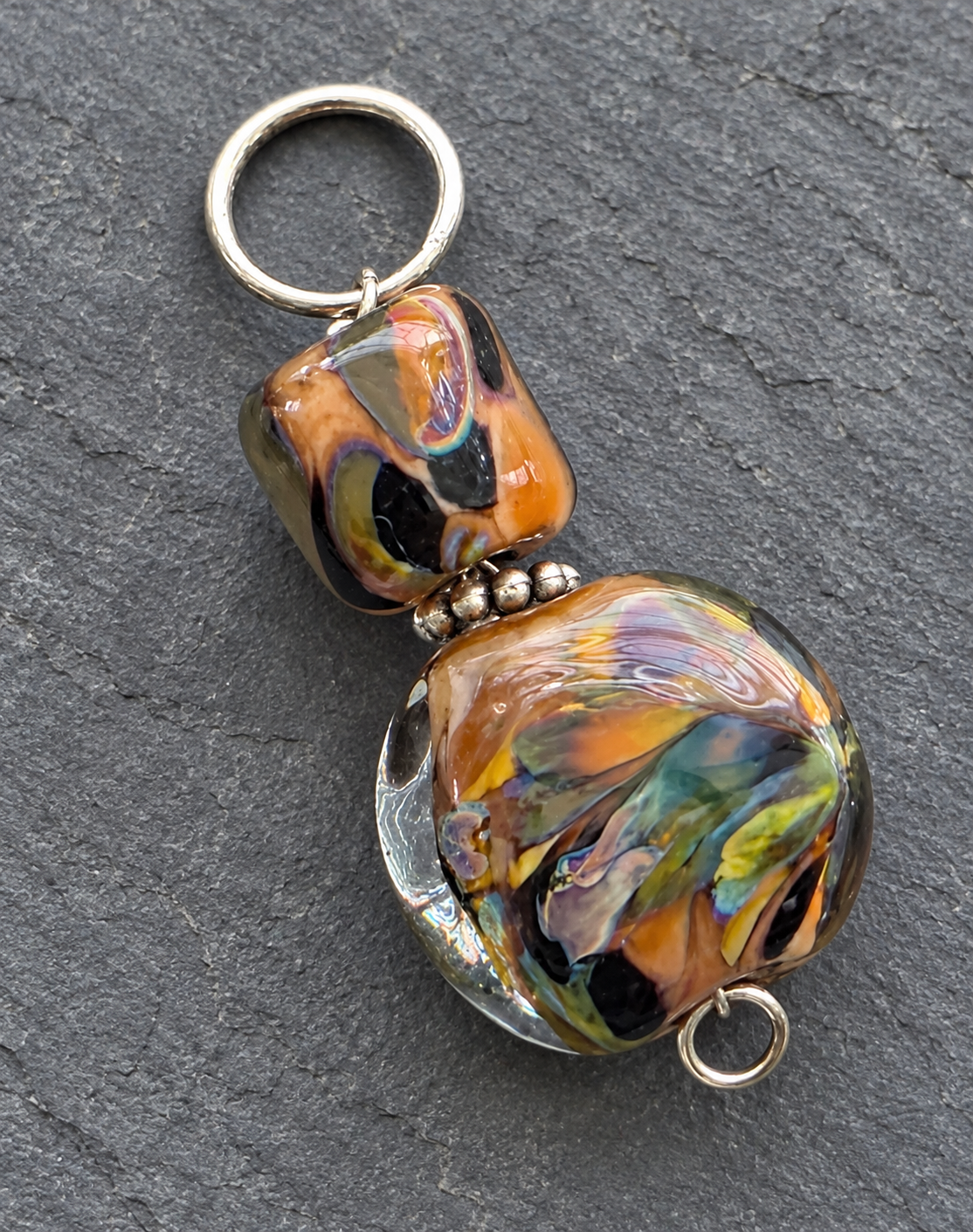 A handmade glass bead keychain featuring a large marbled orange, yellow, green, and black glass bead with a smaller matching square bead, separated by small silver beads, and attached to a silver split ring on a dark gray slate background.