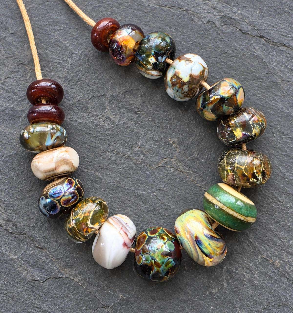 A handmade beaded necklace made of marbled, patterned, and solid glass beads in earthy colors like green, brown, gold, white, and red, strung on a tan cord and displayed on a dark gray slate background.