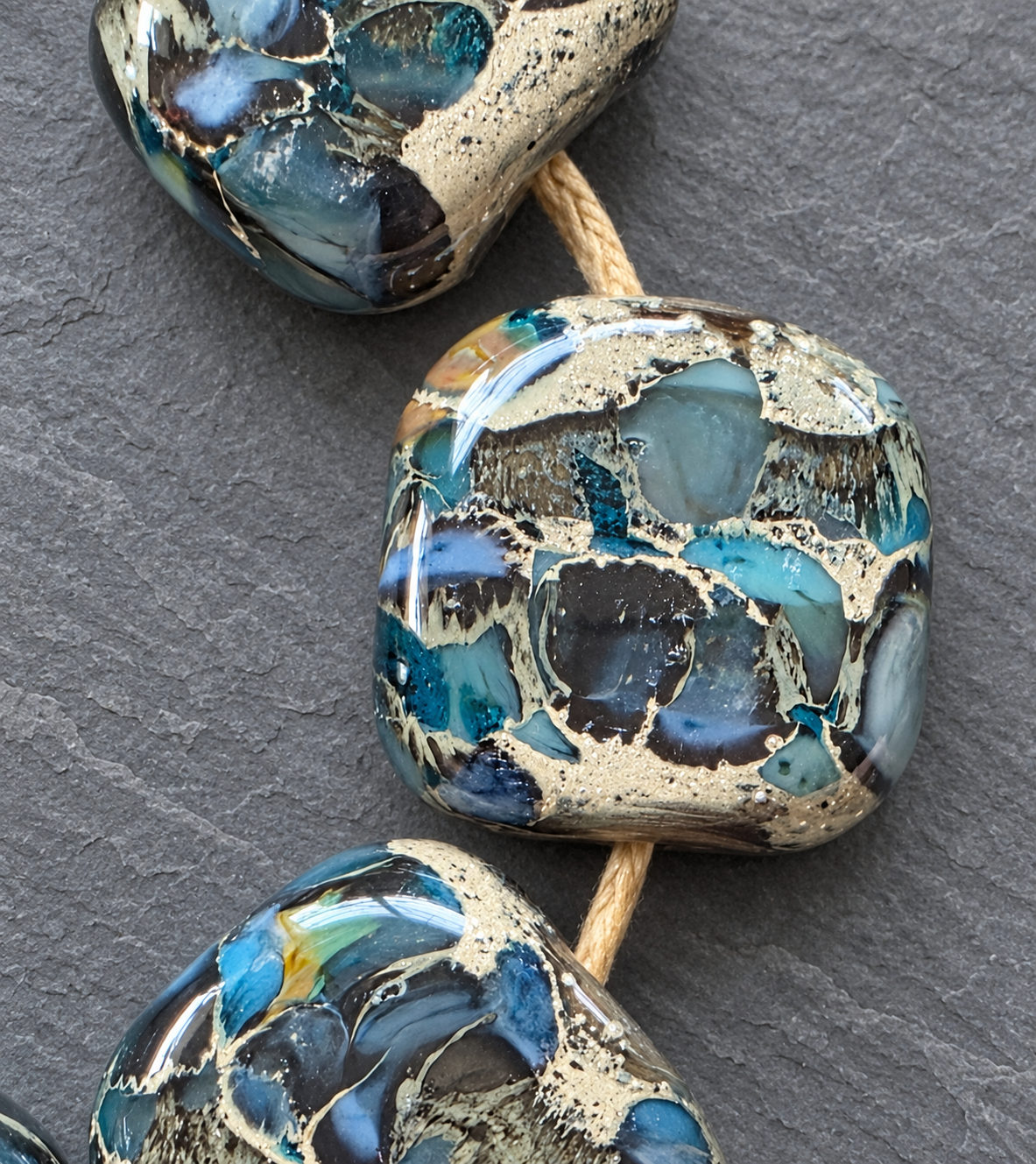 Three polished stone beads with a marbled pattern of turquoise blue, black, and beige, strung on a natural tan cord against a dark gray slate background.