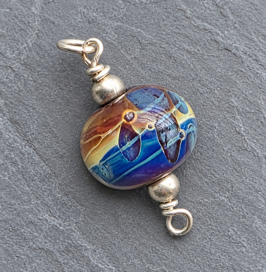 A round glass lampwork bead charm with a colorful swirl pattern of blue, purple, yellow, and brown, finished with silver-toned metal spacers and jump rings on a textured gray slate background.