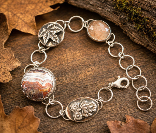A sterling silver bracelet with organic-shaped and engraved charms, displayed on a rustic wooden surface with dried autumn leaves. The bracelet features a round agate stone, a textured silver leaf charm, a faceted peach stone, and floral engravings.